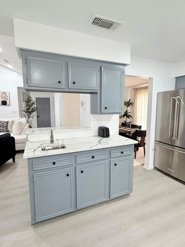 A kitchen with a central island, grey cabinets, a stainless steel refrigerator, and a dining area in the background. - Spring Home near IAH-Houston-Woodlands (Spring)