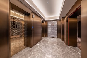 Interior de elevador moderno, com paredes metálicas, piso de mármore e teto com iluminação embutida.