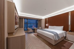Quarto de hotel moderno com uma cama grande, uma escrivaninha, uma cadeira e vista para a cidade à noite.