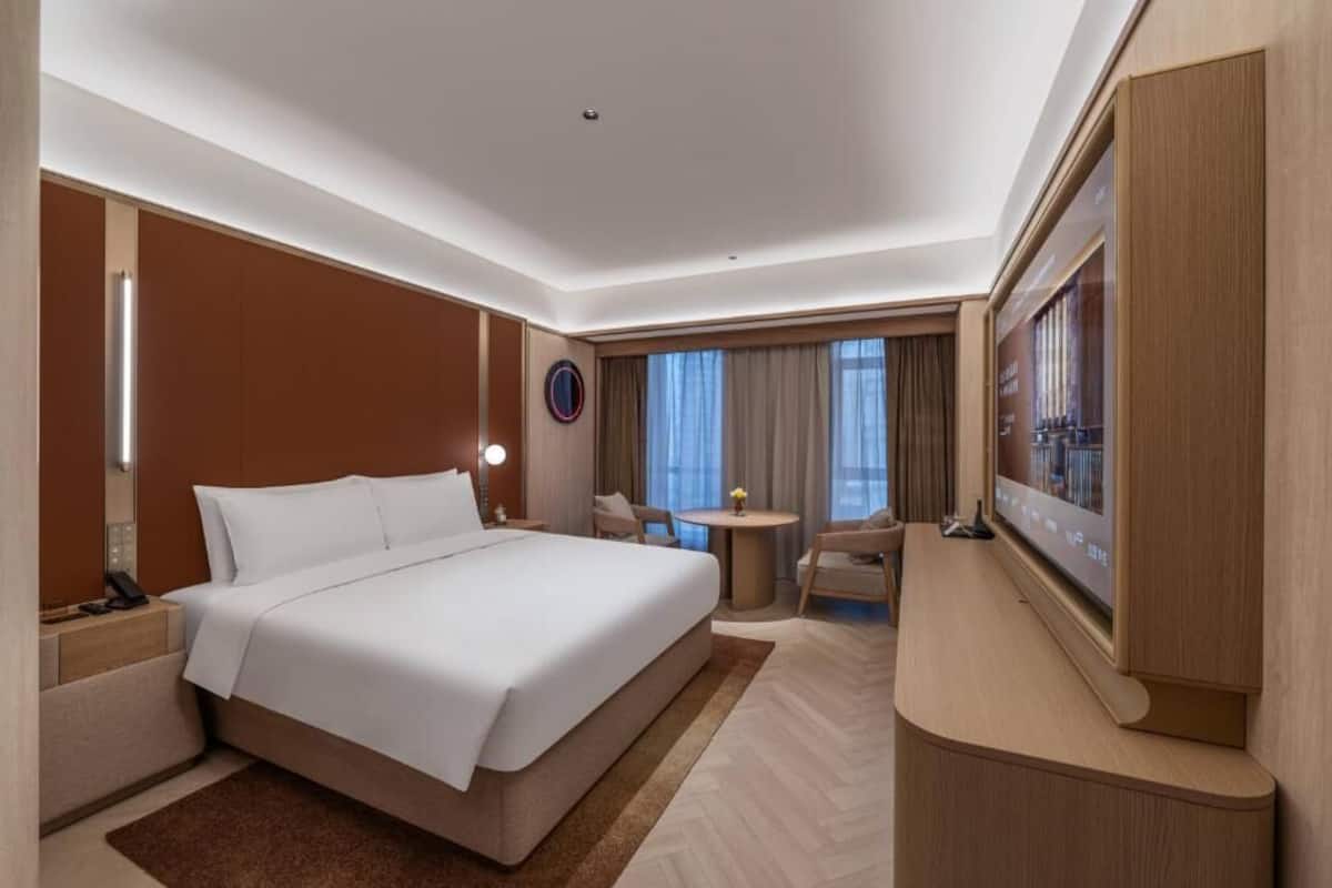 A hotel room with a large bed, a desk with a flat-screen TV, a small round table, and a chair.