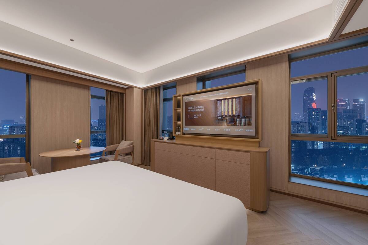 A modern hotel room with a large bed, a desk with a chair, a television, and a city view.