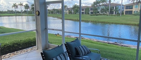 A room with a view of a lake, two chairs with cushions, a table with a vase, and a clock on the wall.