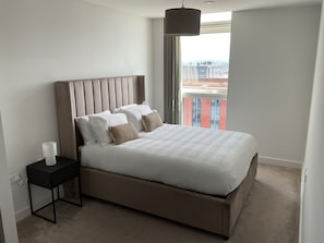 Ein modernes Schlafzimmer mit einem großen Bett, einem Nachttisch, einer Lampe und Blick auf die Stadt durch ein großes Fenster.