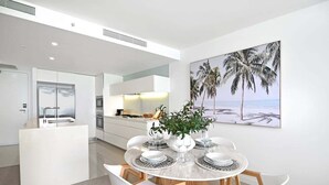 A modern kitchen with white cabinetry, a dining area with a round table and chairs, and a large beach-themed painting on the wall. - Ocean Breeze at Oracle Level 21 (Gold Coast)