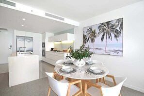 Eine moderne Küche mit weißen Schränken, ein Essbereich mit einem runden Tisch und Stühlen sowie ein großes Gemälde im Strandhaus-Stil an der Wand.