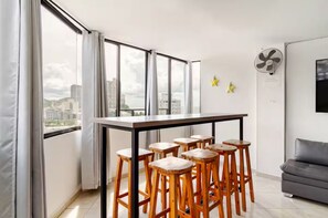 A modern apartment with a dining area featuring a black table and wooden stools, large windows with curtains, a grey sofa, and a wall-mounted fan. - Rodadero Sol & Mar (Gaira)