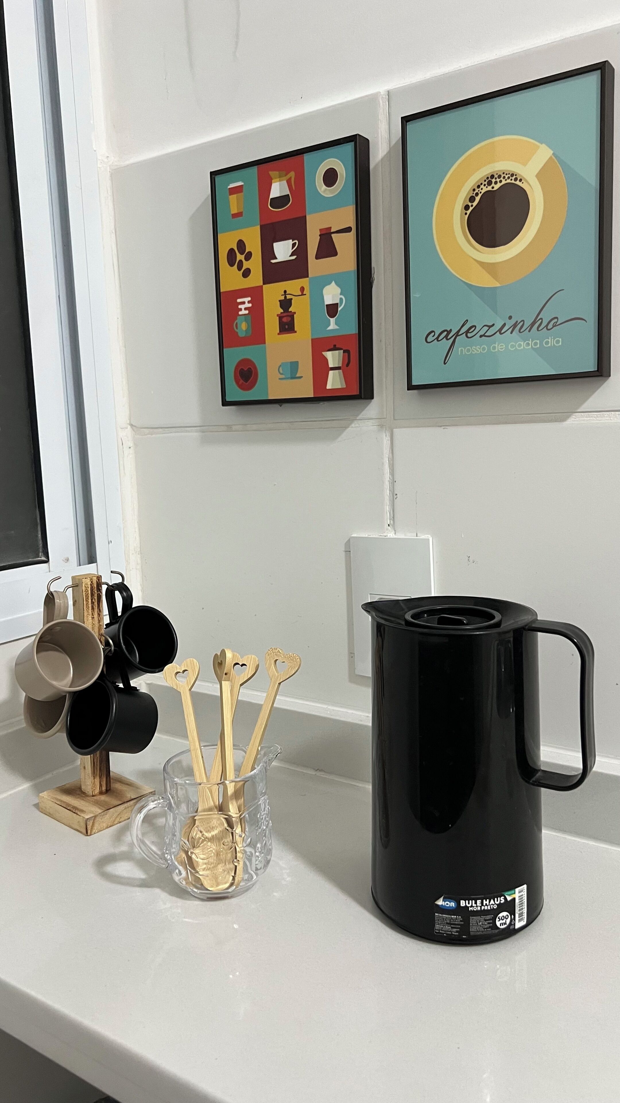 Una encimera de cocina con cafetera, tazas y utensilios. Hay dos cuadros en la pared.
