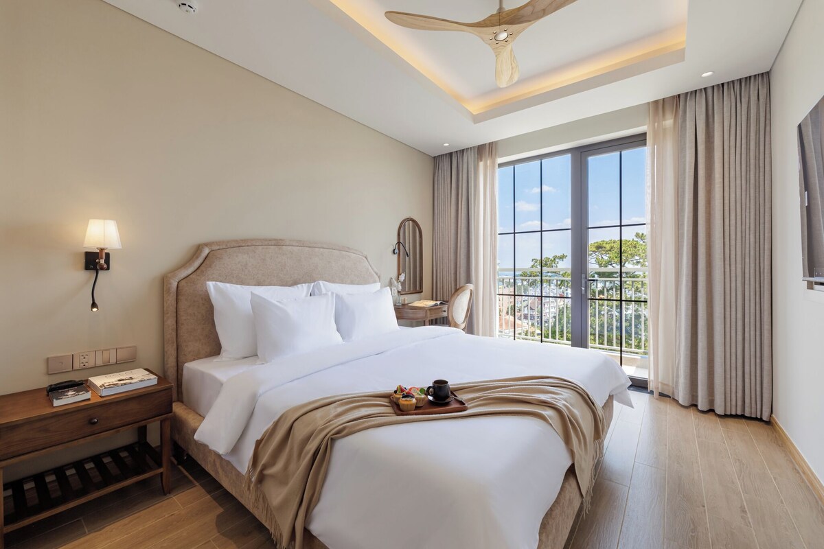 Quarto de hotel com uma cama grande, uma mesa de cabeceira com luminária, um criado-mudo e ventoinha no teto, além de vista para o exterior.