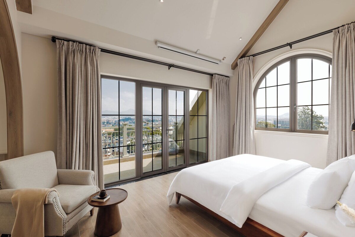Quarto de hotel com uma cama grande, um sofá, uma mesinha e vista para a paisagem urbana através de janelas do chão ao teto.