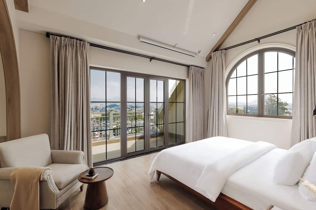 A hotel room with a large bed, a sofa, a small table, and a view of a cityscape through floor-to-ceiling windows.