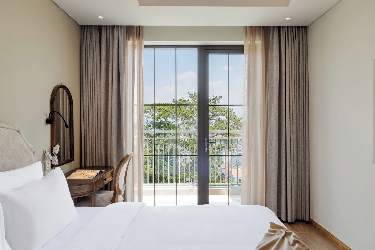 Quarto de hotel com uma cama grande, uma escrivaninha, uma cadeira e vista para uma varanda com cortinas.