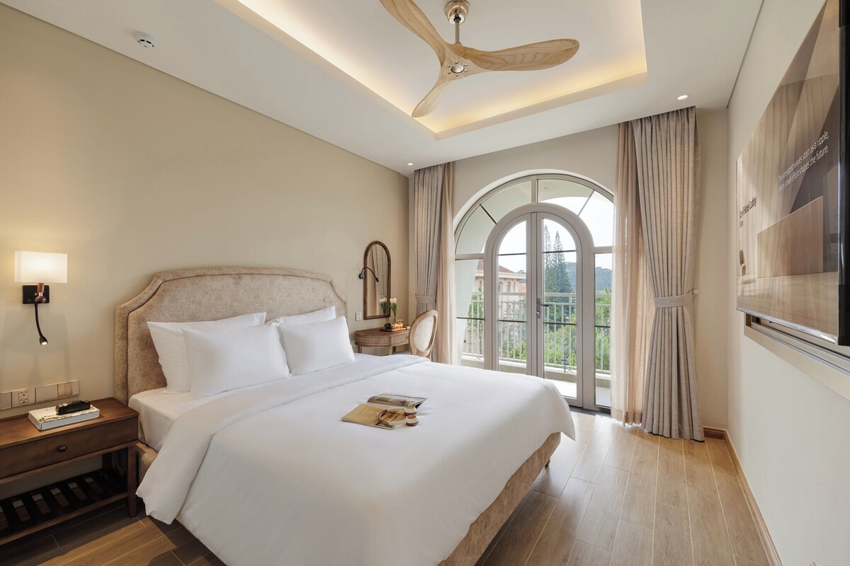 Quarto de hotel com uma cama grande, um ventilador de teto, uma mesa de cabeceira com abajur, uma janela com cortinas e vista para o exterior.
