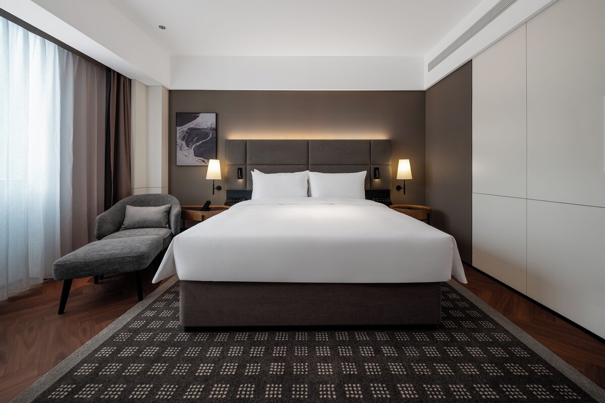Una habitación de hotel moderna con una cama grande, dos lámparas de noche, una silla y una alfombra estampada.