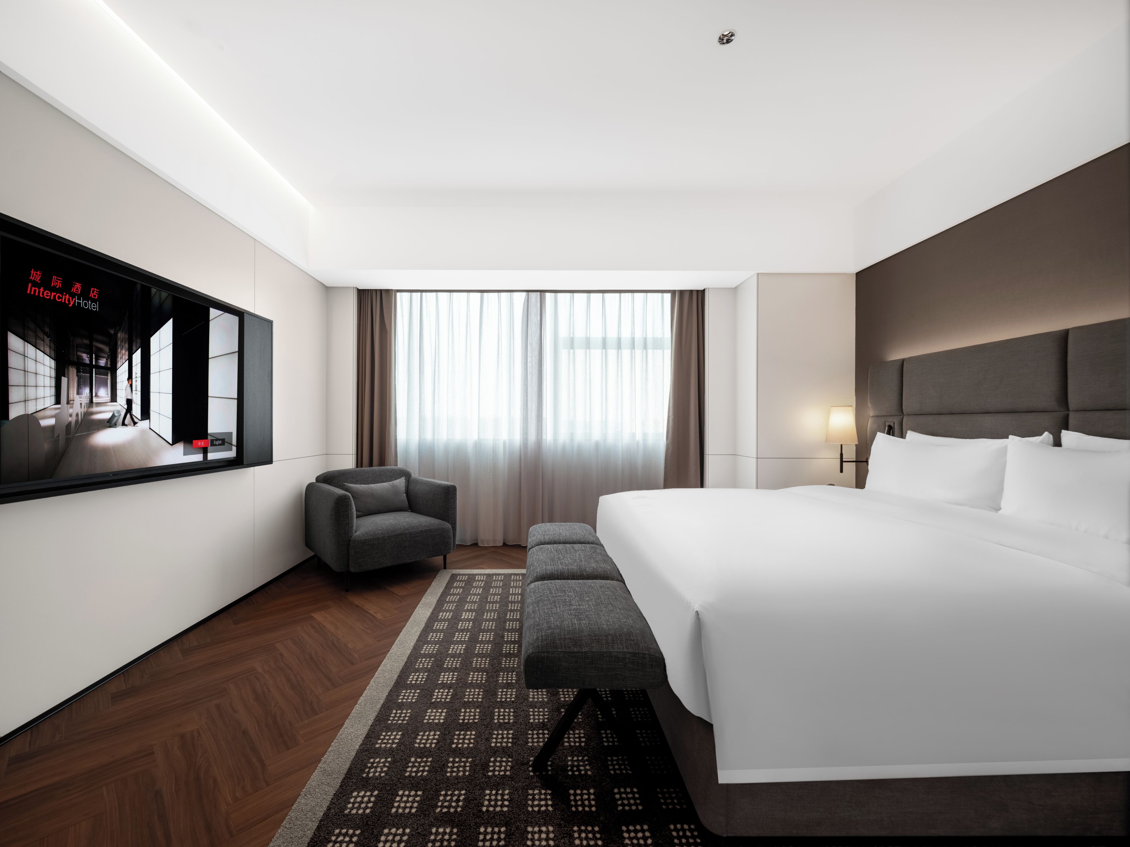 Habitación de hotel con una cama grande, un televisor montado en la pared, una silla, una mesita y una ventana con cortinas.