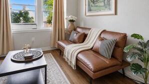 A brown leather sofa with a throw blanket and striped pillows, a round table with a decorative tray, a large plant, and a framed landscape painting on the wall. - Walkerville 1BR | Parking + WiFi + Workspace (Windsor)