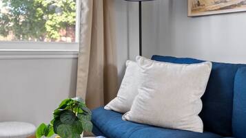 A room with a blue sofa, a round wooden coffee table with a plant and a gramophone, a floor lamp, and a window with curtains.