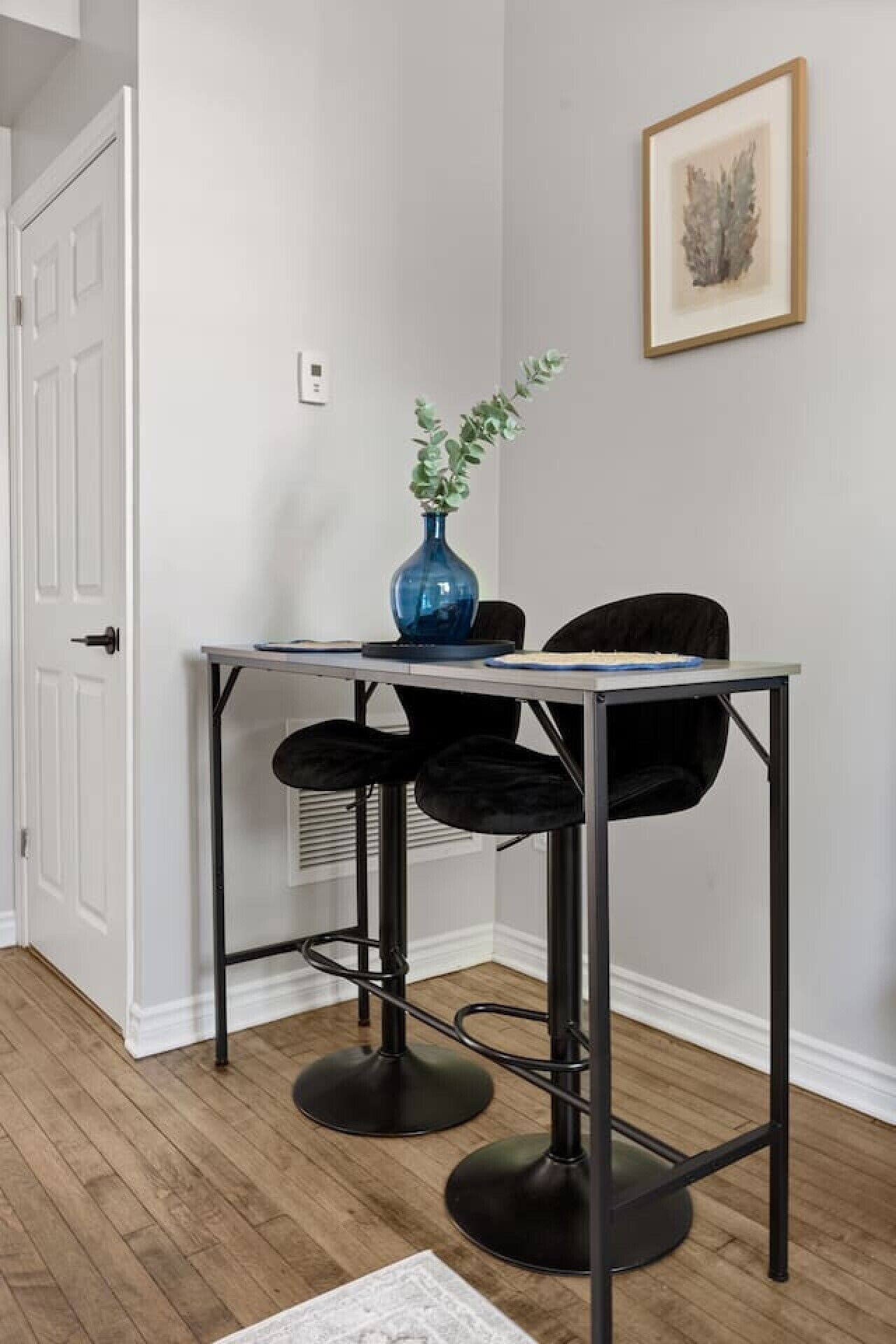 Quarto de canto com porta branca, uma pequena mesa com um vaso e duas cadeiras, e um quadro na parede.