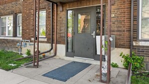Een bakstenen gebouw met een donkere deur, een metalen hekwerk en een blauwe mat bij de ingang.