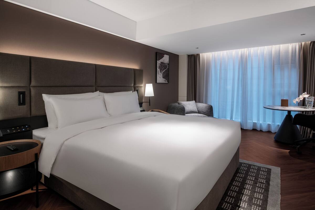 A hotel room with a large bed, a desk with a chair, a small table with a chair, a lamp, and a window with curtains.
