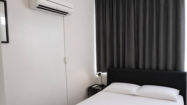Une chambre d’hôtel comprenant un lit, une table de chevet, une lampe, un appareil de climatisation et une fenêtre avec des rideaux.