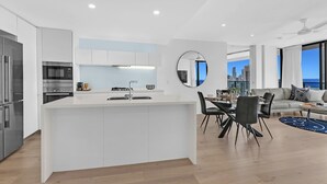 A modern kitchen with a central island, dining area with chairs, and a view of a cityscape through large windows. - Ocean Perfection at Oracle Level 23 (Gold Coast City)