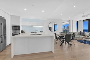 A modern kitchen with a central island, dining area with chairs, and a view of a cityscape through large windows. - Ocean Perfection at Oracle Level 23 (Gold Coast City)