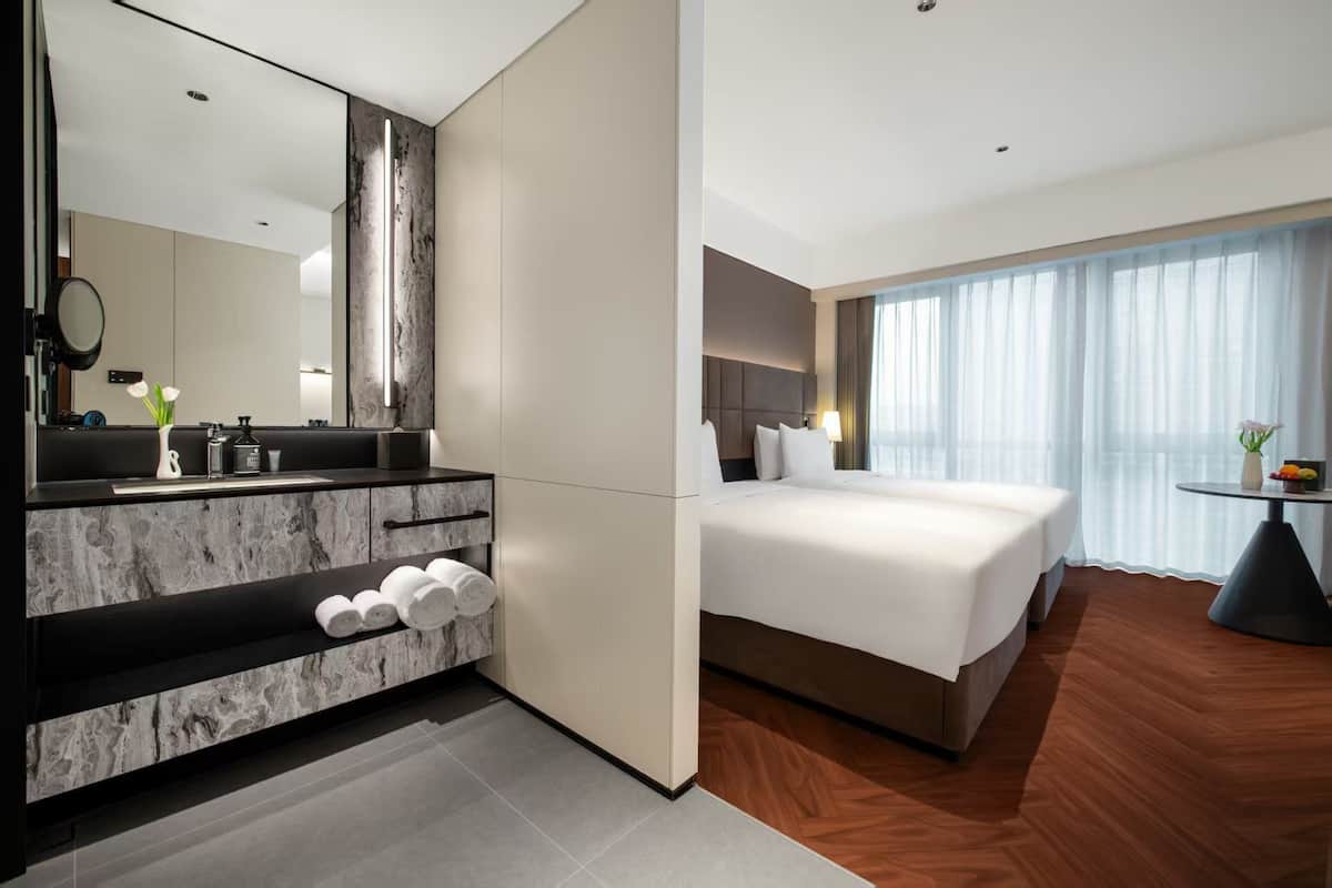 Habitación de hotel moderna con tocador de baño de mármol, una cama grande y vistas de la ciudad a través de cortinas translúcidas.