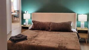 A bedroom with a large bed, two bedside lamps, a chandelier, and a view of a dining area.