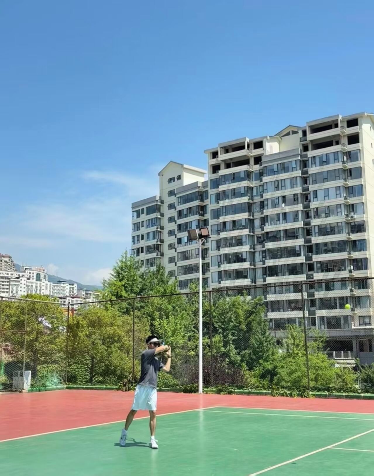 A person playing tennis on an outdoor court with a multi-story building in the background.