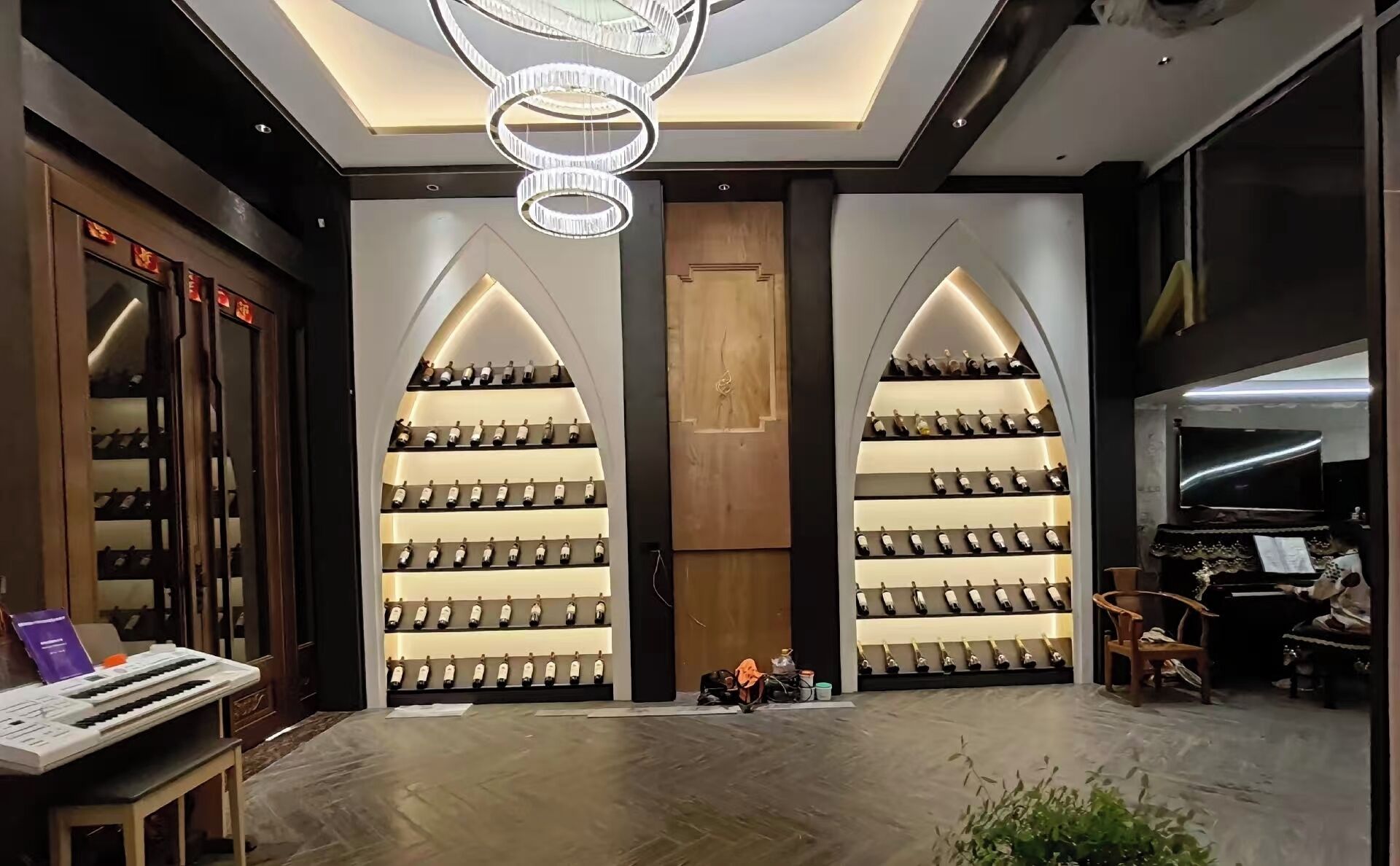 A modern interior with a central wooden door, two symmetrical wine racks on either side, a chandelier, and a piano.