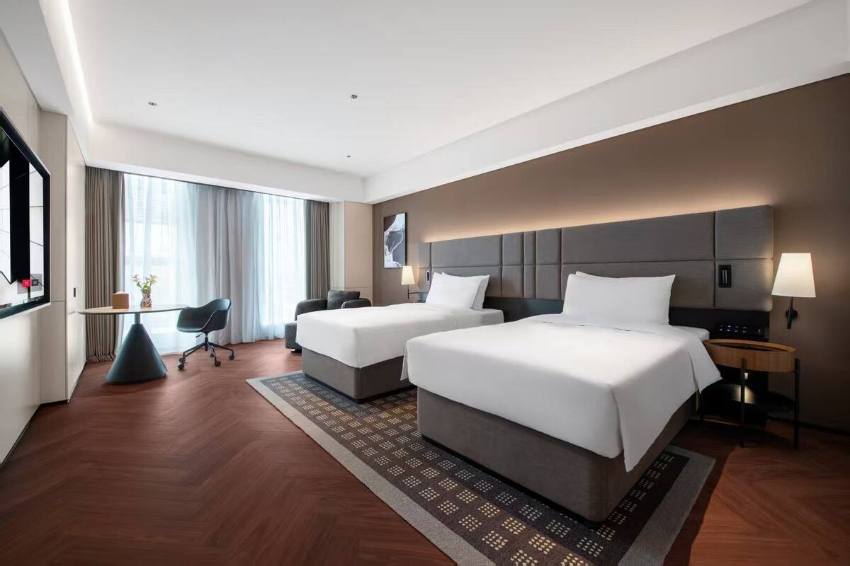 Habitación de hotel con dos camas, un escritorio, una silla y un ventanal con cortinas.