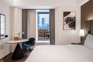 A hotel room with a bed, a desk with a chair, a small table with a vase of flowers, a painting on the wall, and a view of the city through the window.