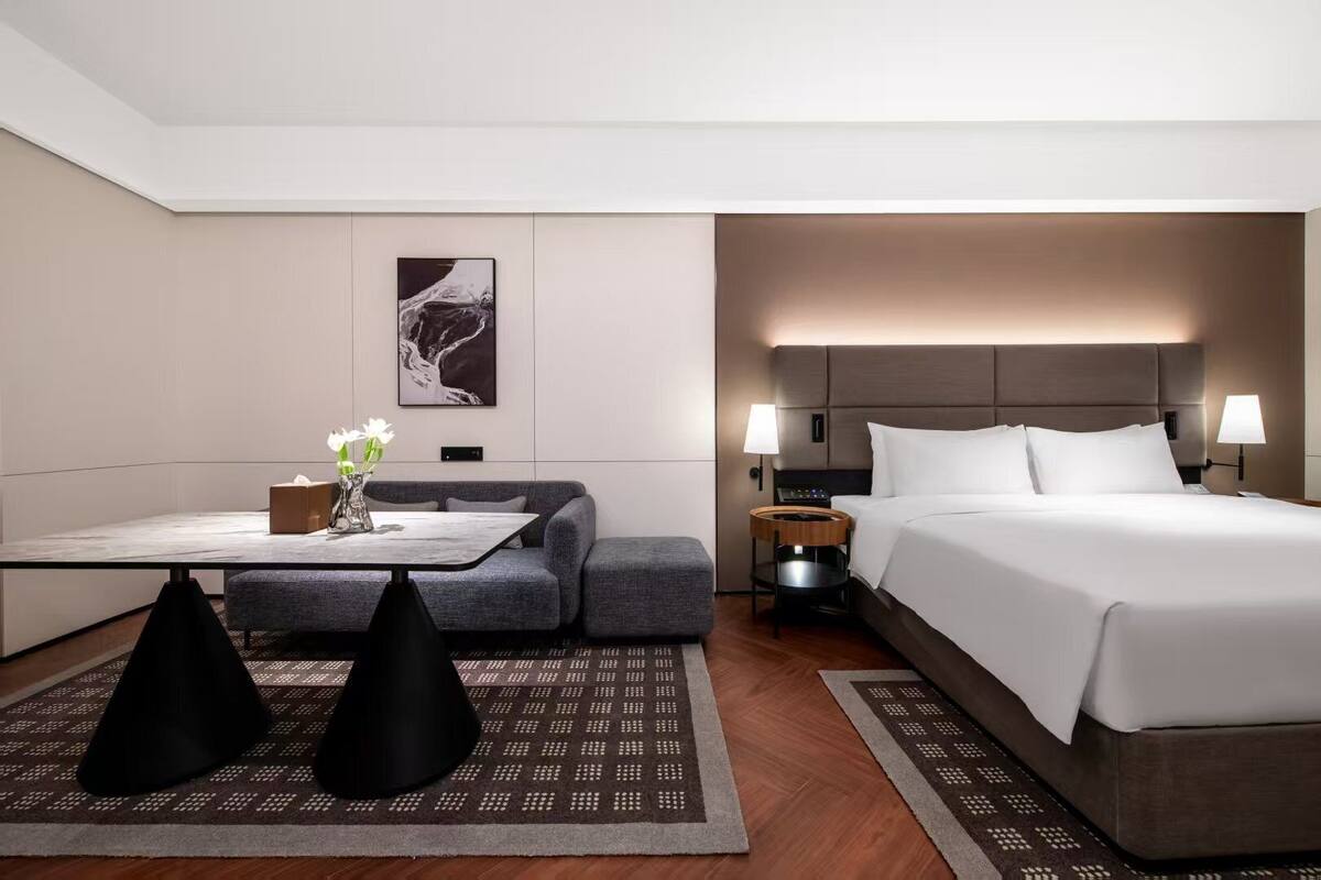 Una habitación de hotel moderna con una cama, mesitas de noche, un sofá, una pequeña mesa con un florero y una obra de arte colgada en la pared.