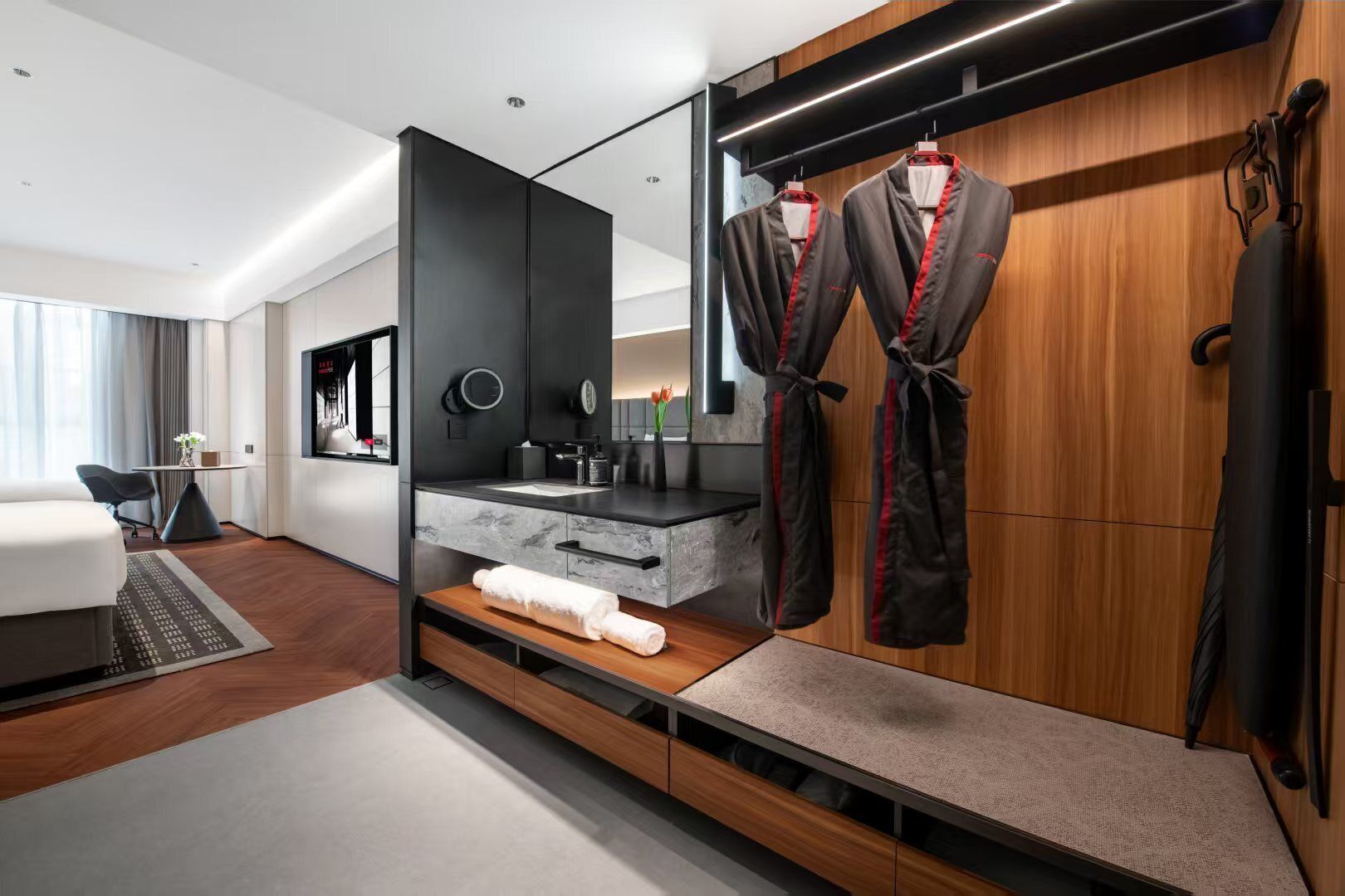 A modern hotel room with a wooden wardrobe, a built-in vanity with a mirror, and a seating area with a small table and chairs.