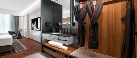 A modern hotel room with a wooden wardrobe, a built-in vanity with a mirror, and a seating area with a small table and chairs.