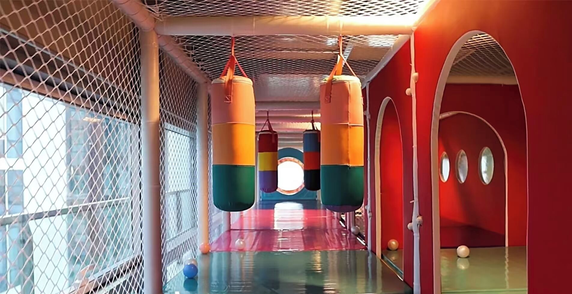 An indoor play area with a netted enclosure, padded punching bags, and a red wall with circular windows.