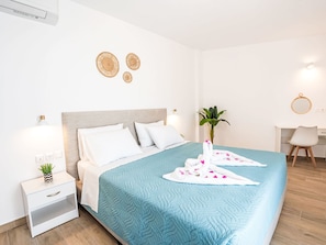 Ein ordentlich bezogenes Bett mit einer türkisfarbenen Steude, weißen Kissen und einem Handtuch mit rosa Blumen. Ein Nachttisch mit Lampe und einer kleinen Pflanze ist ebenfalls vorhanden.