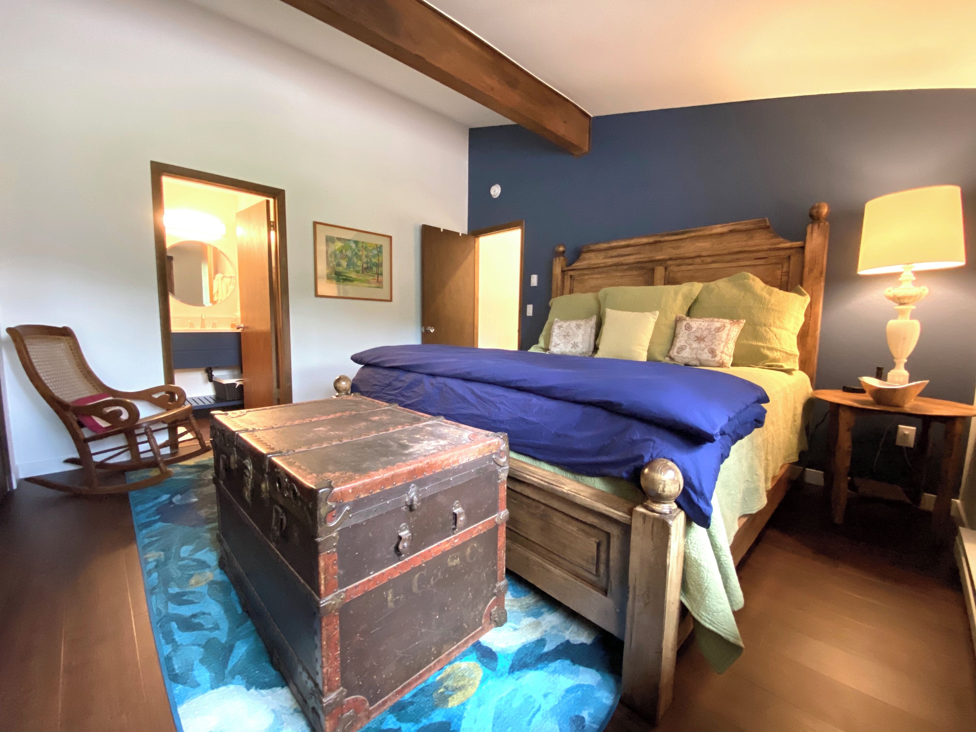 A bedroom with a wooden headboard, a blue bedspread, a rocking chair, a vintage suitcase, and a bedside lamp.