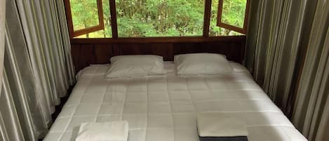 A bed with white bedding, two pillows, and folded blankets in a room with wooden walls and a view of greenery through a window.