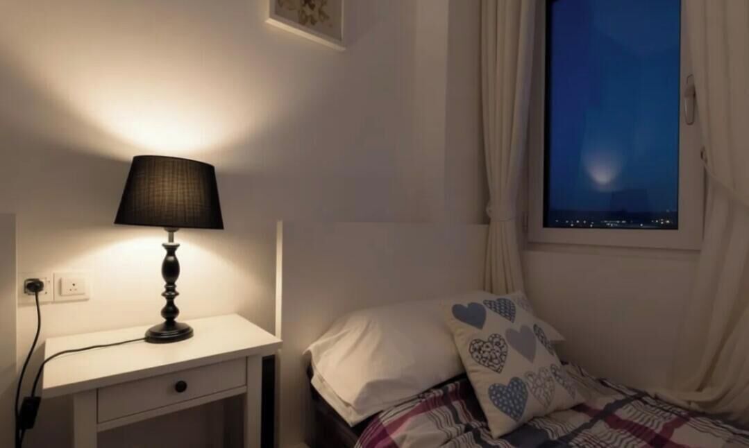 Een slaapkamer met nachtkastje, lamp, bed met hartvormige kussens en een raam met uitzicht op de nachthemel.