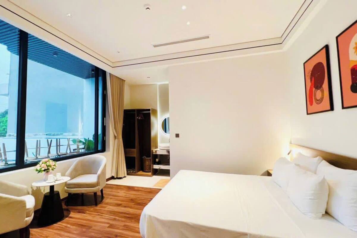 Habitación de hotel con una cama, un área de estar con sillas, una mesita con un florero y ventanas amplias que ofrecen una vista.