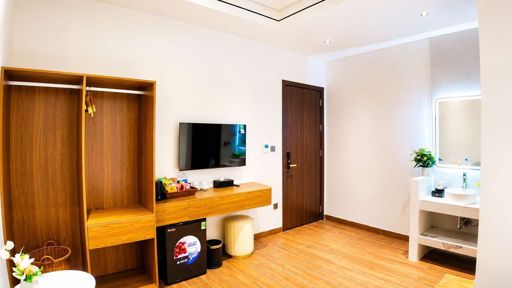 A hotel room with a wooden wardrobe, a flat-screen TV, a small fridge, a sink, and a mirror.