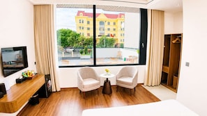 Ett hotellrum med ett stort fönster, två fåtöljer, ett litet bord, en tv och ett skrivbord i trä.