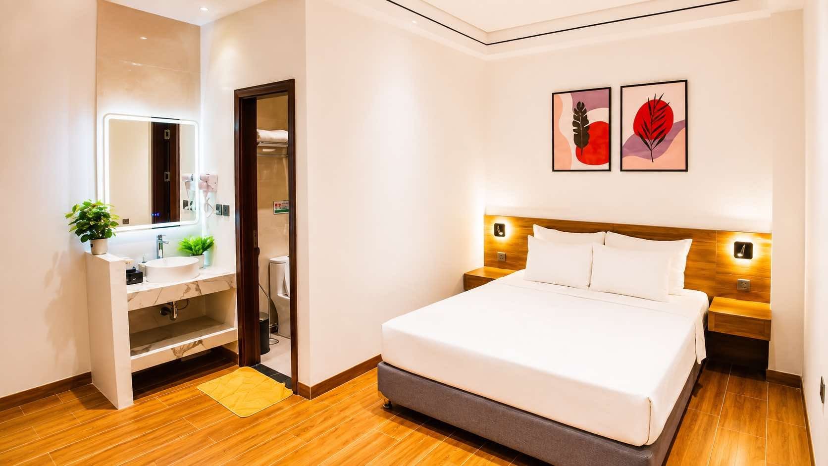 Habitación de hotel con cama, mesitas de noche, espejo, lavamanos y una planta en maceta.