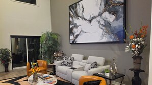 A modern living room with a grey sofa, orange armchairs, a coffee table with magazines, a large abstract painting, and a potted plant. - Lenexa Executive Getaway (Lenexa)
