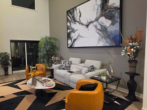 Ein modernes Wohnzimmer mit einem grauen Sofa, orangefarbenen Sesseln, einem Couchtisch mit Zeitschriften, einem großen abstrakten Gemälde und einer Topfpflanze.