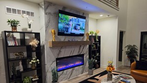 A modern living room with a fireplace, a flat-screen TV mounted above it, and a bookshelf filled with books and decorative items. - Lenexa Executive Getaway (Lenexa)