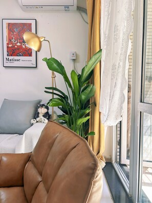 Une pièce avec un canapé en cuir marron, une plante verte, une œuvre d’art encadrée au mur et une fenêtre avec des rideaux.