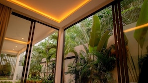 Quarto moderno com amplas janelas, uma cama e plantas tropicais visíveis do lado de fora.