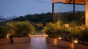 Uma área externa moderna com deck de madeira, plantas em vasos e um toldo de vidro.
